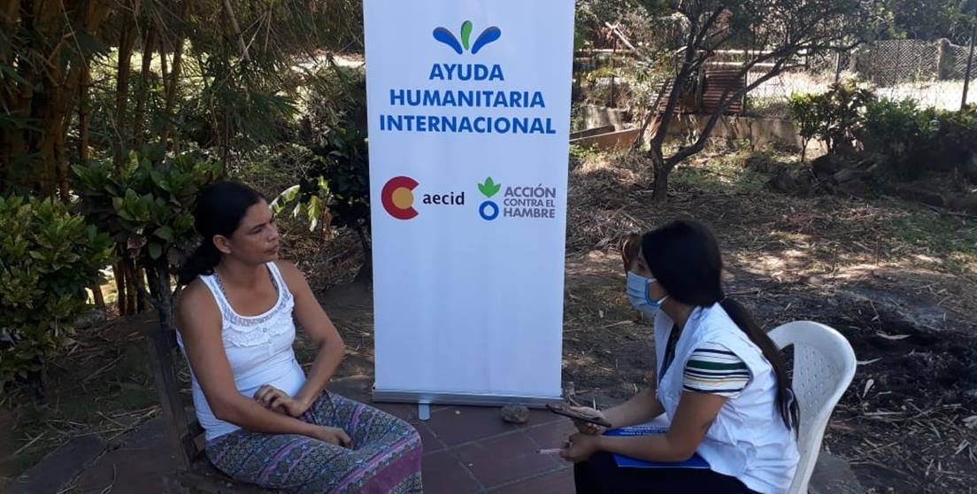 Dos mujeres charlando sentadas con cartel de Ayuda Humanitaria Internacional de fondo